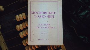 A vintage Moscow guidebook — "Moskovskie Tolkuchki" (Brief Guide, 1993) — rests on a worn abacus and dark leather, photographed from above in soft, moody light. The faded paper, purple typography and subtle stains tell a quiet story of markets, travel and everyday life, while the textured beads and cracked cover evoke tactile nostalgia and the charm of analog objects.